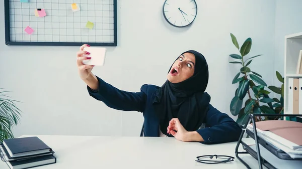 Mujer de negocios musulmana haciendo muecas mientras toma selfie en el teléfono inteligente en la oficina - foto de stock