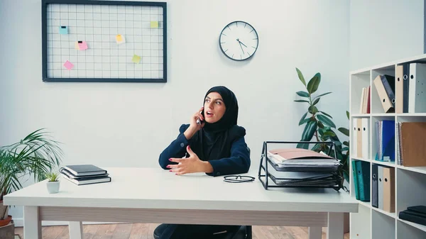 Muslim businesswoman in hijab sitting at desk and talking on smartphone in office — Stock Photo