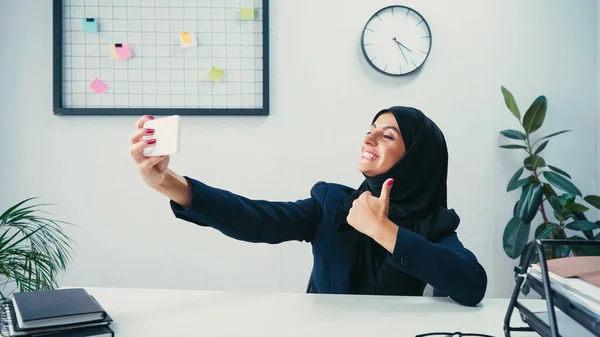 Happy muslim businesswoman showing thumb up while taking selfie on smartphone in office — Stock Photo