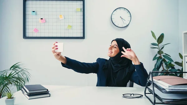 Feliz musulmana mujer de negocios mostrando signo de paz y tomar selfie en el teléfono inteligente en la oficina - foto de stock