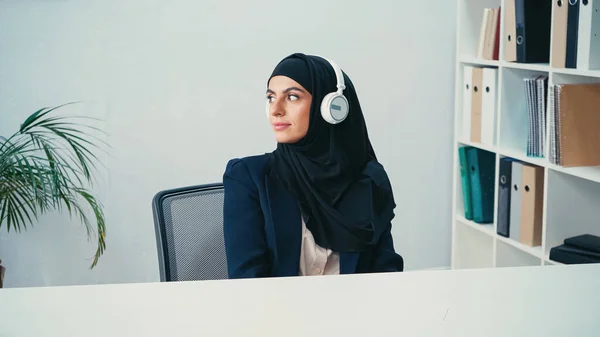 Sonriente mujer de negocios árabe en hijab escuchando música en auriculares inalámbricos en la oficina - foto de stock