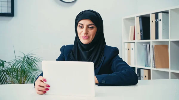 Femme musulmane souriante dans le hijab en regardant la tablette numérique au bureau — Photo de stock