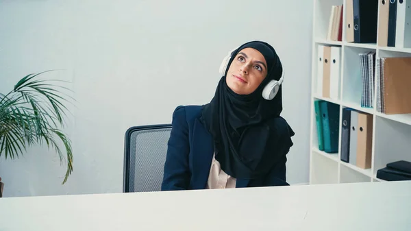 Dreamy arabian businesswoman in hijab listening music in headphones in office — Stock Photo