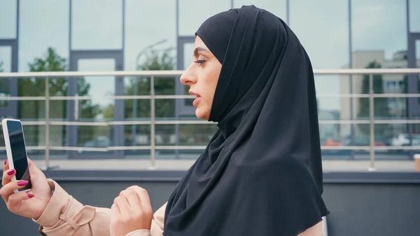 Vista lateral da jovem mulher muçulmana ter chamada de vídeo na rua urbana — Fotografia de Stock