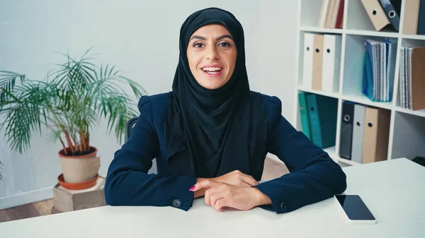Jovem mulher muçulmana sorrindo perto do smartphone com tela em branco na mesa — Fotografia de Stock