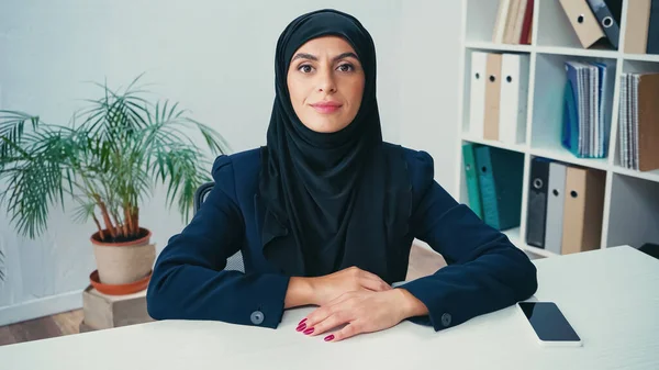Jovem mulher muçulmana perto de smartphone com tela em branco na mesa — Fotografia de Stock