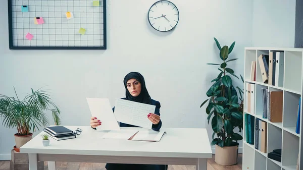 Junge muslimische Frau im Hidschab schaut sich Dokumente an und sitzt am Schreibtisch im Büro — Stockfoto