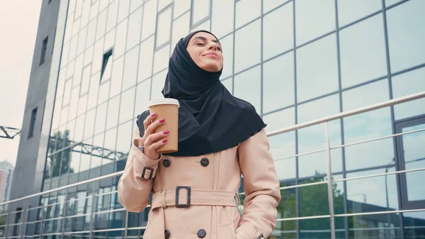 Vista de ángulo bajo de la mujer musulmana complacida en hijab y gabardina sosteniendo taza de papel cerca del edificio exterior - foto de stock