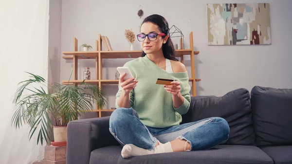 Young woman using smartphone and credit card on couch — Stock Photo