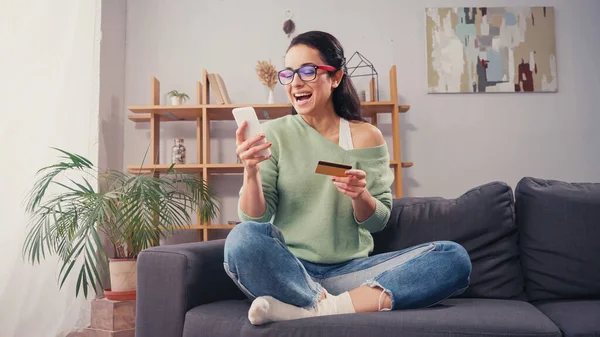 Excited woman in eyeglasses using smartphone and credit card — Stock Photo