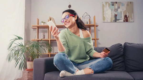 Pleased woman holding credit card and using smartphone at home — Stock Photo