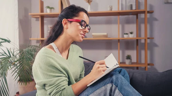 Joven estudiante de anteojos escribiendo en cuaderno en casa — Stock Photo