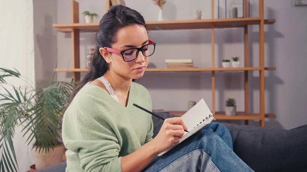 Estudiante morena en anteojos escribiendo en cuaderno en sala de estar - foto de stock