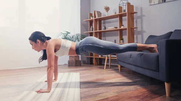 Vista lateral de la deportista haciendo tablón en sofá y colchoneta de fitness - foto de stock