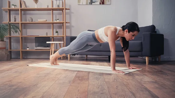 Vue latérale de la sportive tendue debout dans la planche sur tapis de fitness à la maison — Photo de stock