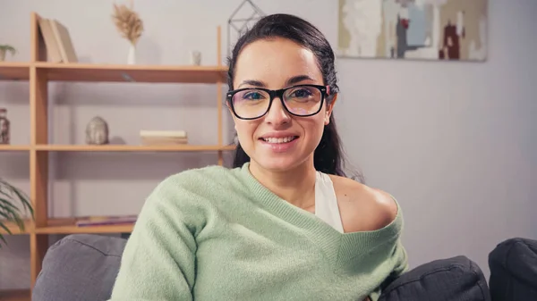 Mujer joven en gafas de vista sonriendo a la cámara en el sofá - foto de stock