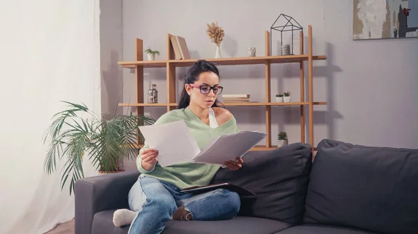 Student in eyeglasses looking at papers while sitting on couch — Stock Photo