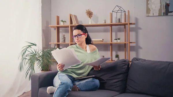 Estudiante concentrado en anteojos mirando documentos en sofá en sala de estar — Stock Photo