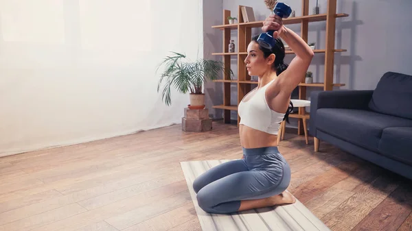 Vue latérale de l'entraînement de la sportive brune avec haltère sur tapis de fitness rayé à la maison — Photo de stock