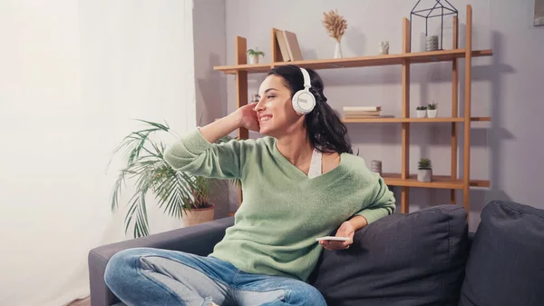 Cheerful woman in headphones holding smartphone on couch — Stock Photo