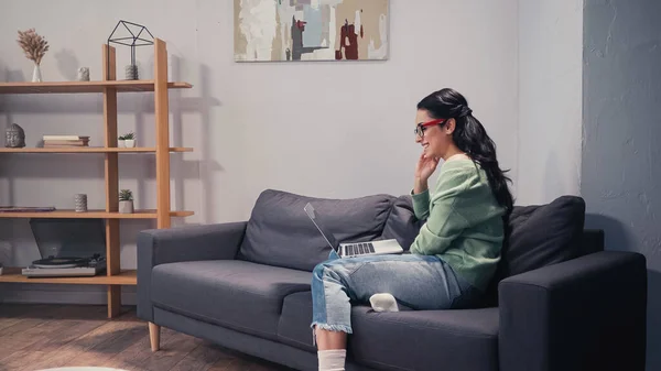Seitenansicht einer positiven Frau mit Brille, die auf dem Sofa auf den Laptop schaut — Stockfoto