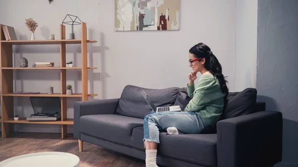 Seitenansicht eines hübschen Telearbeiters in Brille mit Laptop auf der Couch — Stockfoto