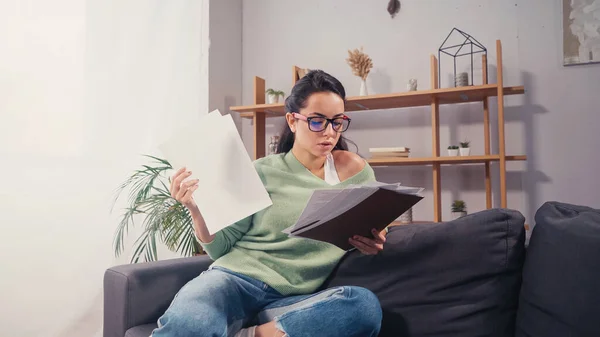 Young brunette student in eyeglasses looking at documents in living room — Stock Photo