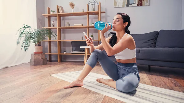 Sportlerin trinkt Wasser und benutzt Handy auf Fitnessmatte im Wohnzimmer — Stockfoto