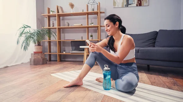 Mulher positiva em sportswear usando smartphone perto de garrafa de esportes em casa — Fotografia de Stock