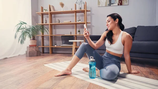 Sportswoman sorrindo ter chamada de vídeo no smartphone perto de garrafa de esportes e tapete de fitness — Fotografia de Stock