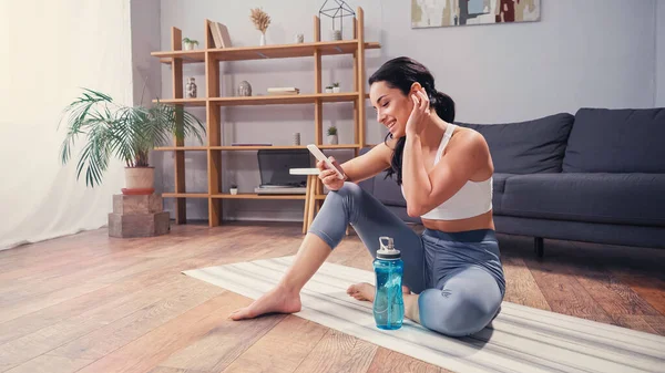 Вид сбоку женщины, которая ведет видеочат на смартфоне, сидя на фитнес-коврике рядом со спортивной бутылкой дома — стоковое фото