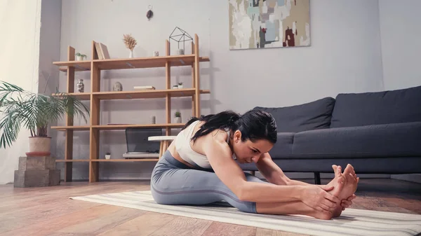 Sportiva in forma toccando le gambe mentre si estende sul tappeto fitness in soggiorno — Foto stock