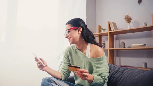 Mujer positiva usando tarjeta de crédito y teléfono inteligente en casa - foto de stock