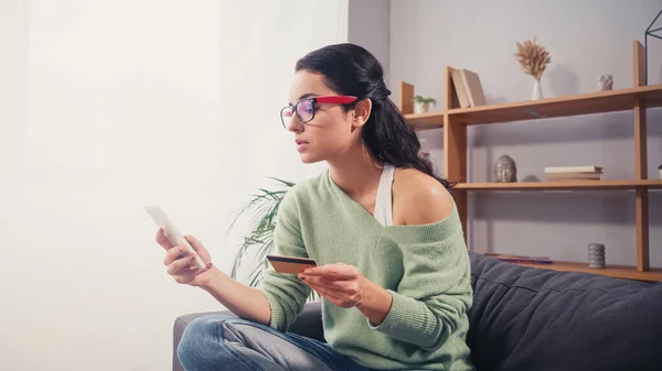 Mujer joven con gafas de vista usando smartphone y tarjeta de crédito durante las compras en línea en casa - foto de stock