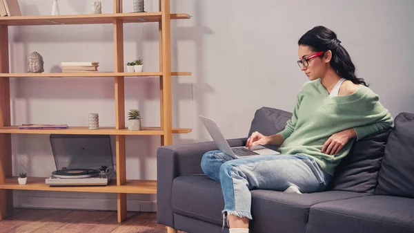 Вид сбоку женщины в повседневной одежде и очках с ноутбуком на диване — стоковое фото