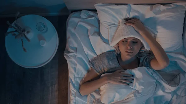 Top view of sick woman with towel on head lying on pillow in bedroom — Stock Photo