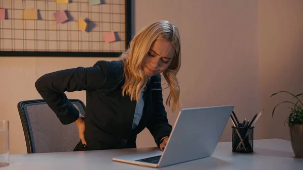 Empresária sofrendo de dor nas costas perto de laptop no escritório à noite — Fotografia de Stock