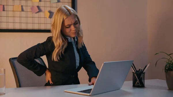 Empresária exausta sentindo dor nas costas perto do laptop no escritório à noite — Fotografia de Stock