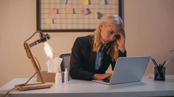 Femme d'affaires épuisée utilisant un ordinateur portable près de pilules, serviette et pulvérisation nasale au bureau — Photo de stock