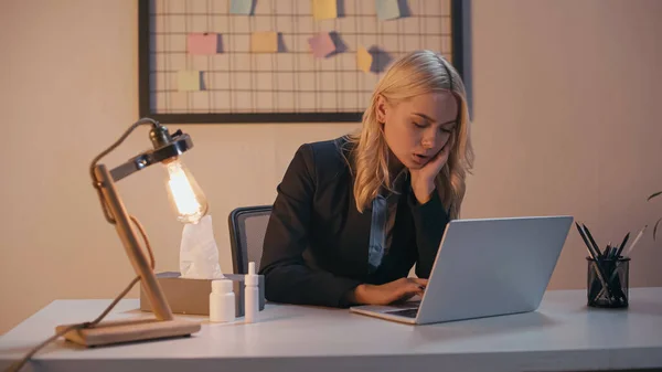 Femme d'affaires malade travaillant sur un ordinateur portable près de pulvérisation nasale, pilules et serviettes au bureau — Photo de stock