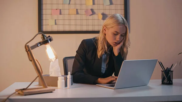 Sick businesswoman working on laptop near nasal spray, pills and napkins in office — Stock Photo