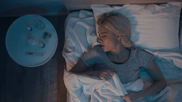 Vista superior de la mujer joven durmiendo cerca del termómetro y pastillas en la mesita de noche - foto de stock