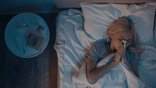 Vue de dessus de la femme avec le nez qui coule couché sur le lit près des pilules sur la table de chevet — Photo de stock