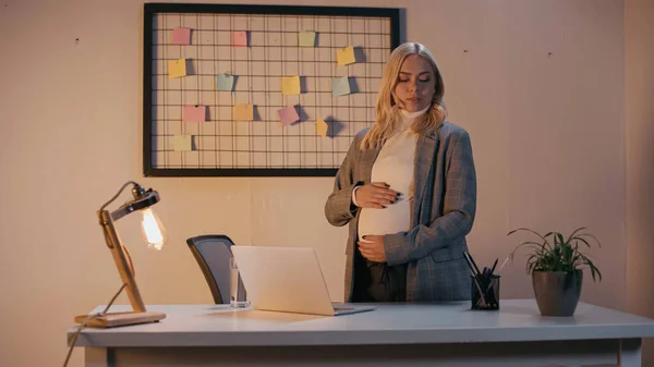 Беременная деловая женщина, стоящая у канцелярских принадлежностей и ноутбука в офисе вечером — стоковое фото