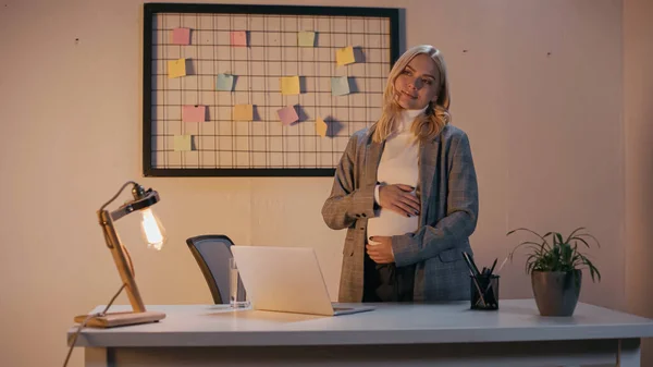Donna d'affari incinta in piedi vicino a computer portatile e vetro d'acqua in ufficio in sera — Foto stock