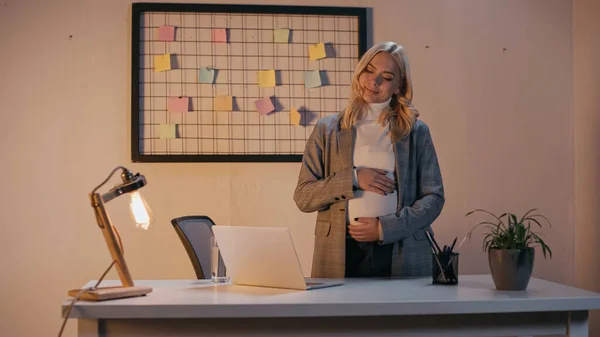Schwangere Geschäftsfrau steht abends neben Laptop im Büro — Stockfoto