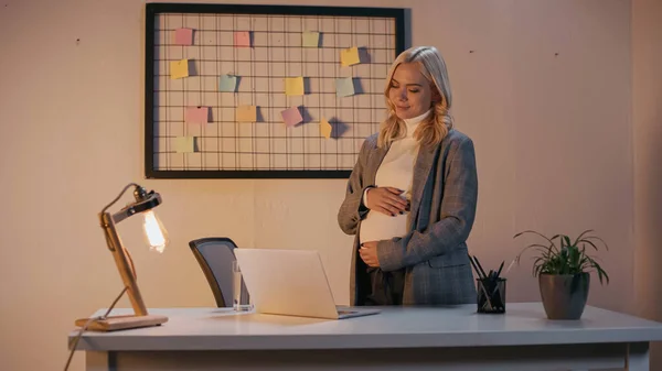 Donna d'affari incinta che tocca la pancia vicino a computer portatile e vetro d'acqua in ufficio in sera — Stock Photo