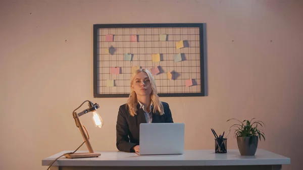 Femme d'affaires regardant la caméra près de l'ordinateur portable et la lampe sur la table de bureau — Photo de stock