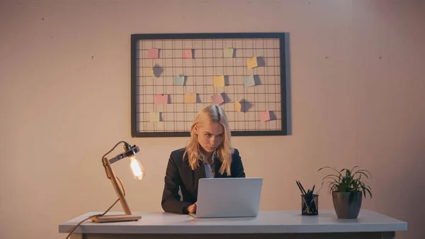 Businesswoman using laptop near lamp and stationery in office in evening — Stock Photo