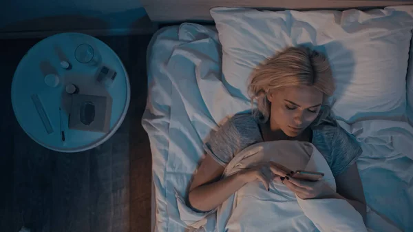 Vue du dessus de la jeune femme utilisant un smartphone près des pilules et un thermomètre électronique sur le lit — Photo de stock